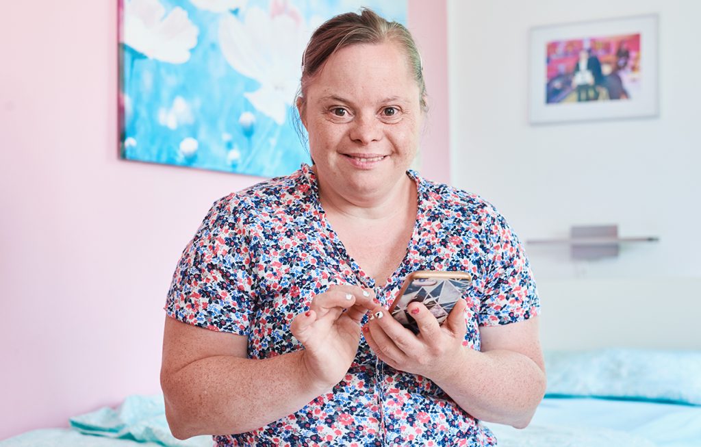 Bild einer geistig behinderten Frau in ihrem Zimmer auf dem Bett sitzend.